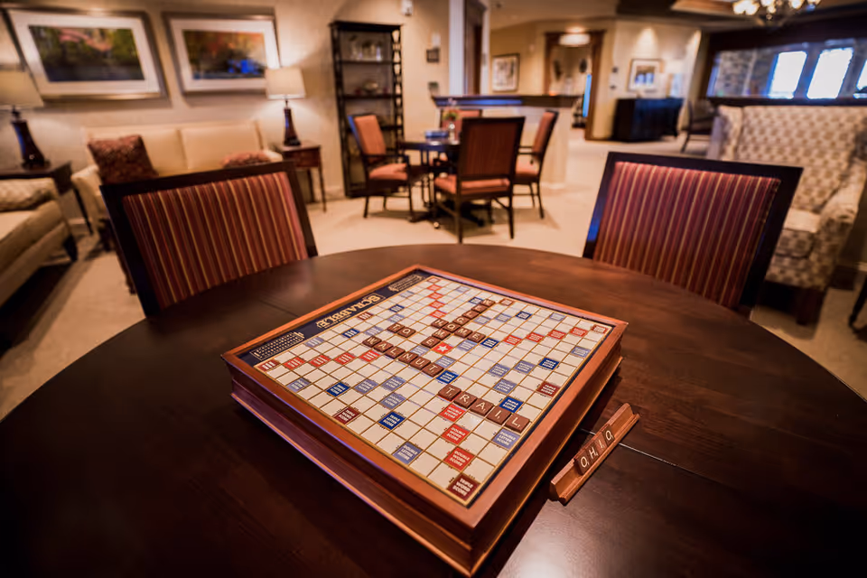 A cozy common area with a dark wooden table in the foreground featuring a Scrabble game in progress with the words THE INN AT WALNUT TRAIL spelled out. The room has several upholstered chairs with striped fabric, a beige sofa, framed artwork on the walls, and warm lighting from table lamps. Additional seating and tables are visible in the background, creating a comfortable and inviting atmosphere.