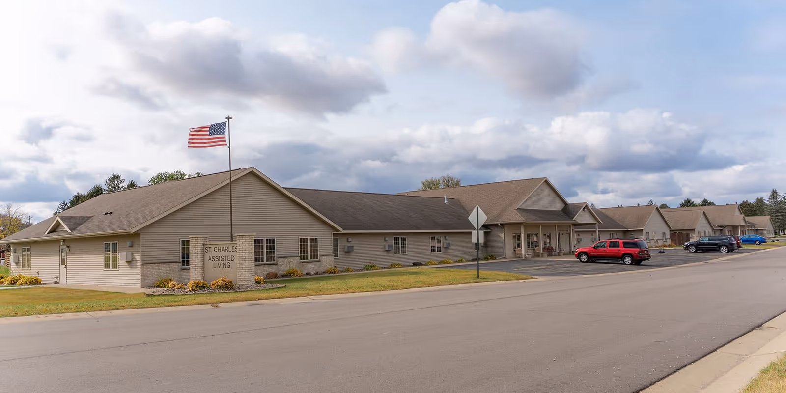 Exterior view of St. Charles Assisted Living facility showing a single-story building with beige siding and a pitched roof. An American flag is flying on a flagpole near the building's sign. Several cars are parked in the parking lot under a partly cloudy sky.