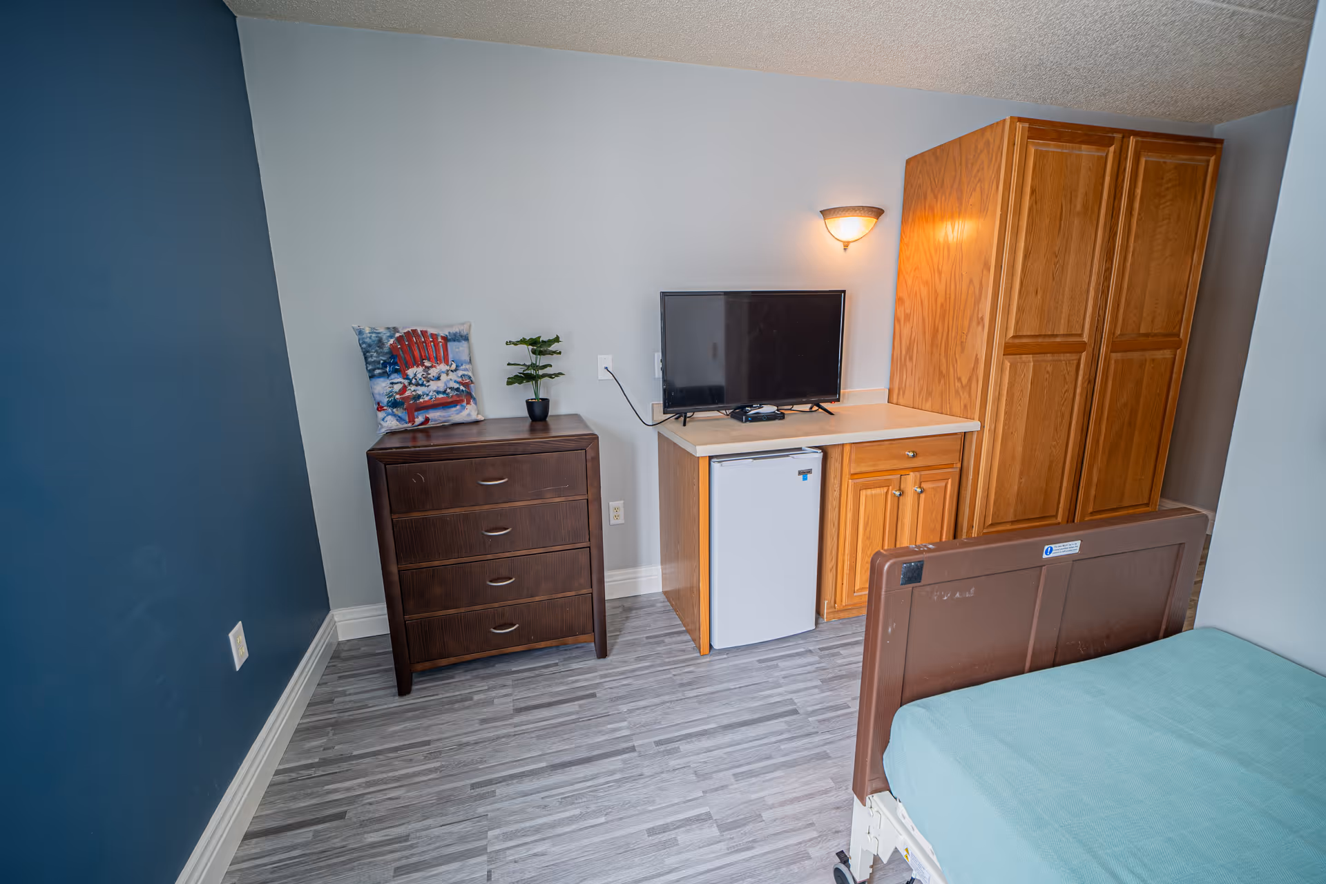 A small room with a hospital-style bed with a teal blanket, a wooden dresser with a decorative pillow and small plant on top, a countertop with a small refrigerator underneath, a flat-screen TV on the counter, and a wooden wardrobe. The walls are painted blue and light gray, and the floor has gray wood-like vinyl flooring.