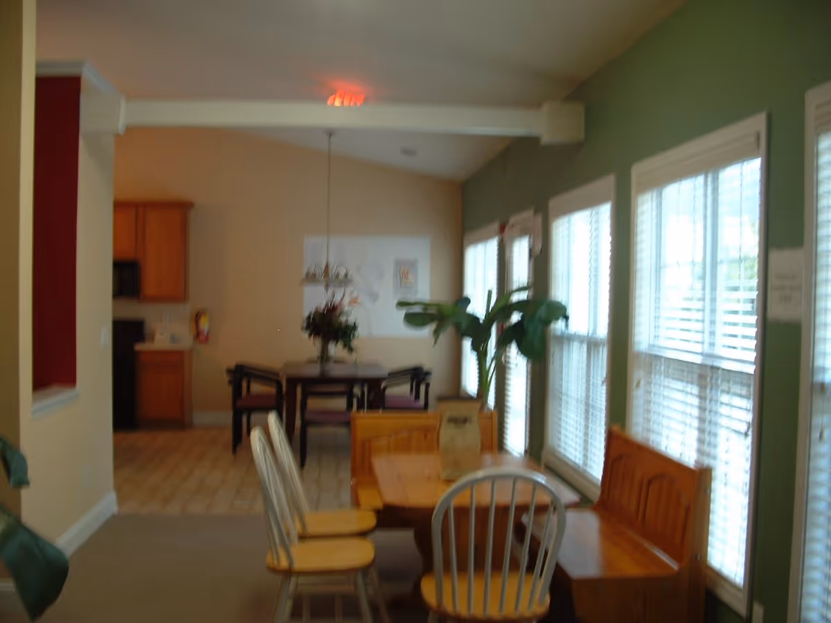 Interior view of a senior living facility dining area with wooden tables and chairs. There are large windows with white blinds on the right side, a green wall, and a potted plant near the windows. In the background, there is another dining table with chairs and a hanging light fixture, and a kitchen area with wooden cabinets.