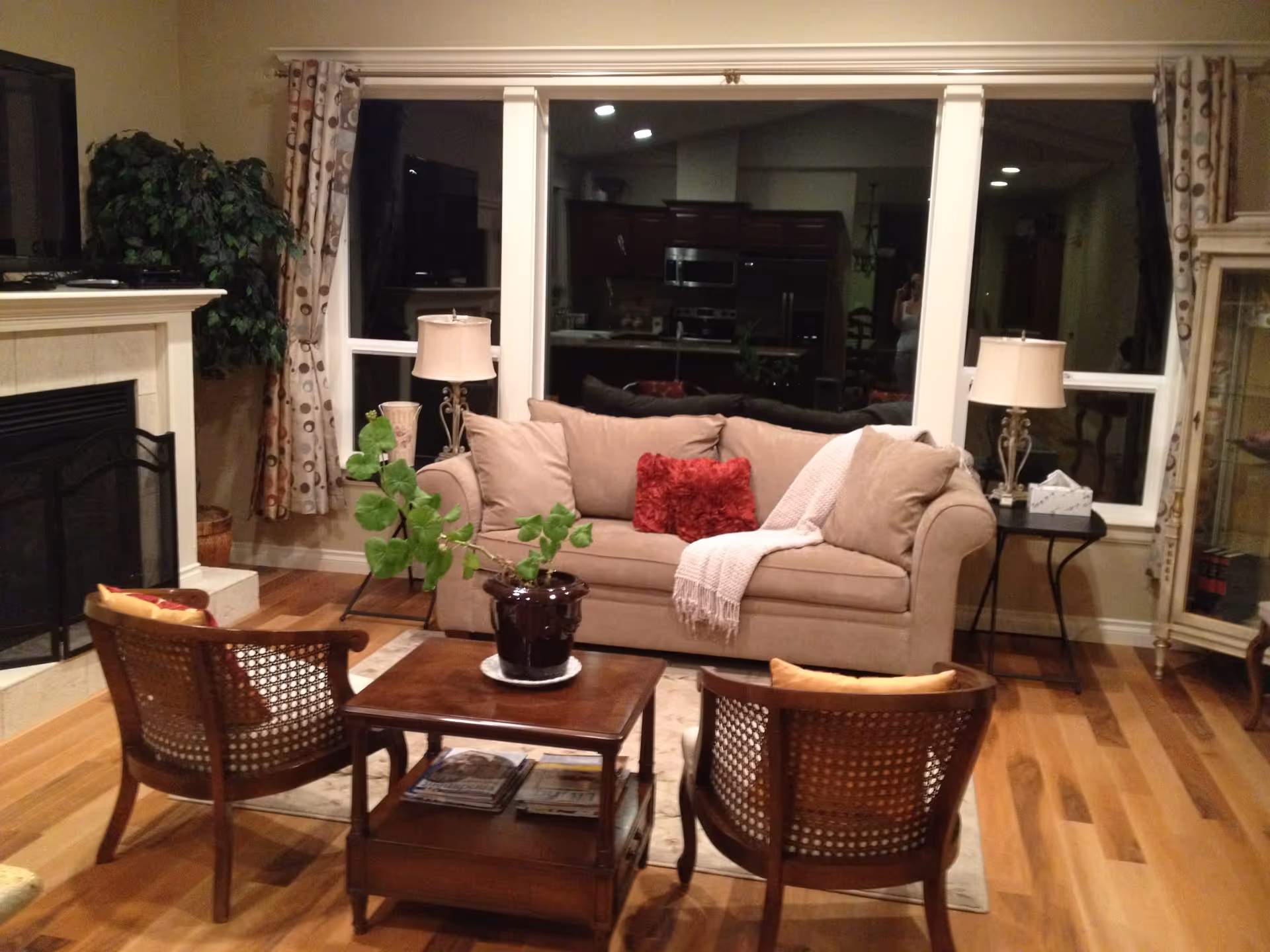 A cozy living room with a beige sofa adorned with a red pillow and a light pink throw blanket. In front of the sofa is a wooden coffee table with a potted plant and magazines underneath. Two wooden chairs with woven backs face the sofa. The room has hardwood flooring, a fireplace on the left, and large windows with patterned curtains. Two table lamps sit on side tables flanking the sofa.