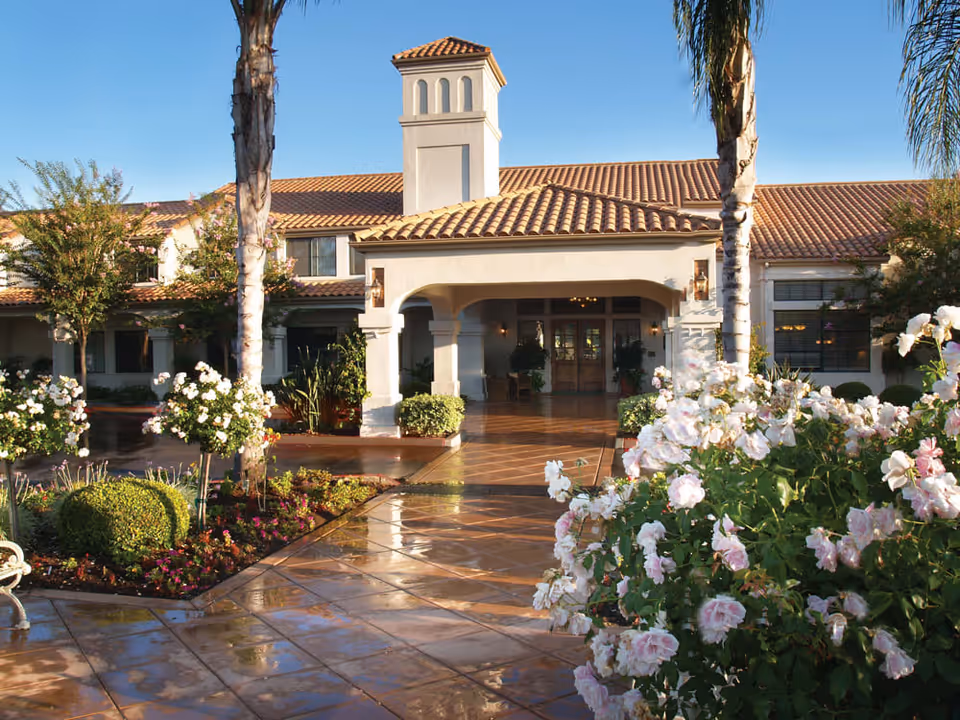 Front exterior view of Oakmont of Escondido Hills facility with a tiled walkway, palm trees, flowering bushes, and a building with a terracotta tiled roof under a clear blue sky.