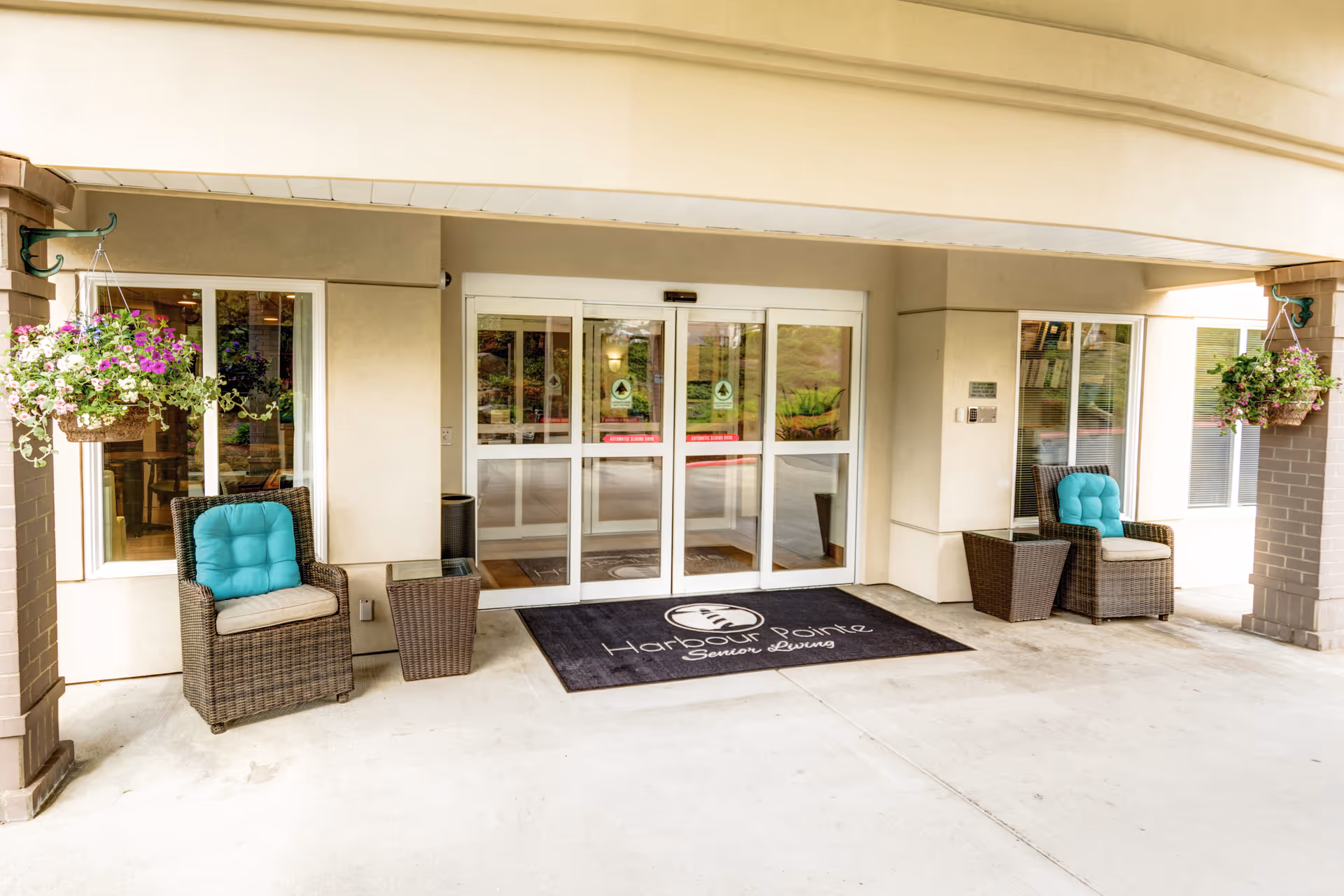 Covered entrance to Harbour Pointe Senior Living with glass automatic doors, a welcome mat bearing the facility name, wicker chairs with teal cushions, and hanging flower baskets.