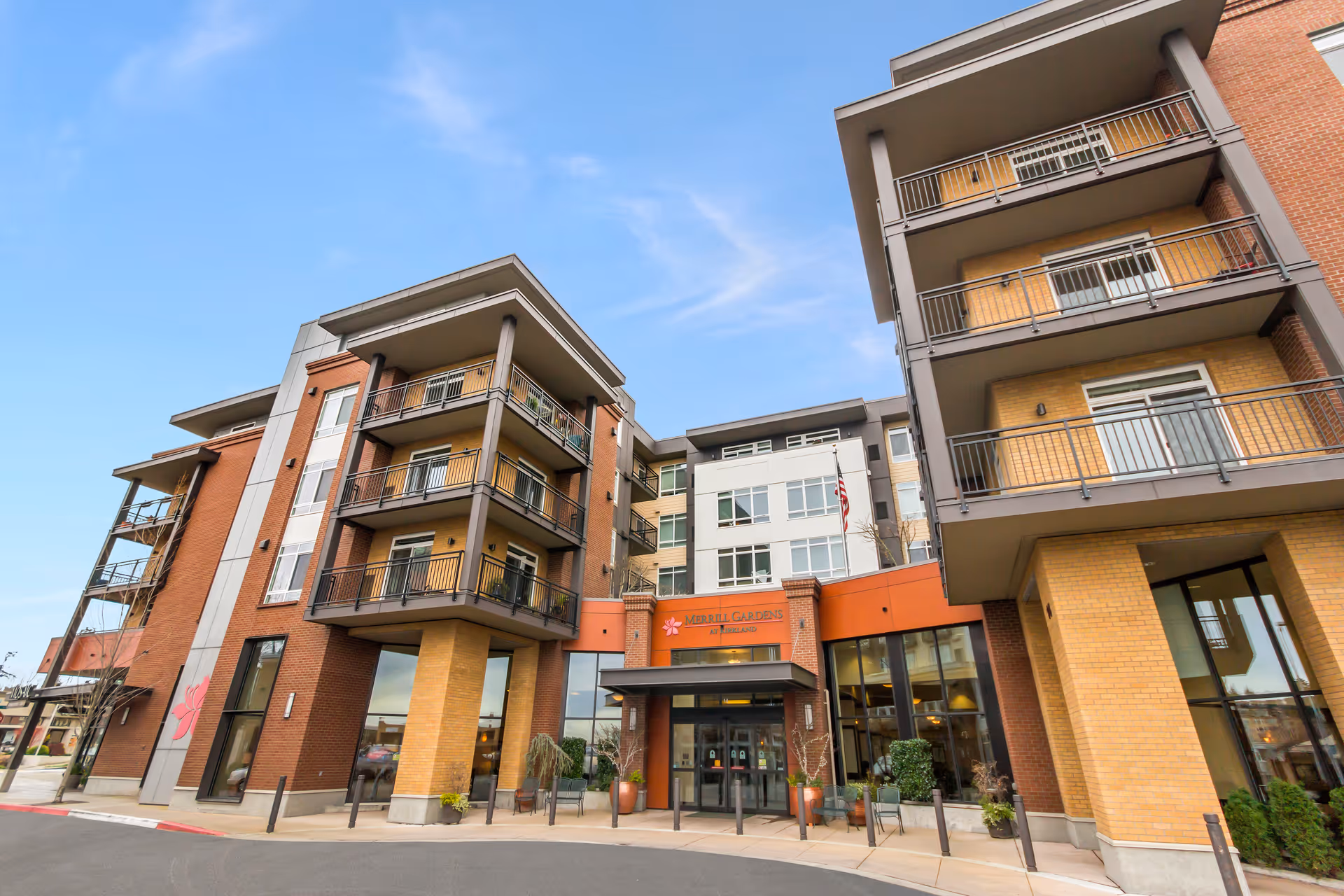 Exterior view of a modern multi-story senior living facility named Merrill Gardens at Kirkland, featuring balconies, large windows, and an entrance with glass doors under a clear blue sky.