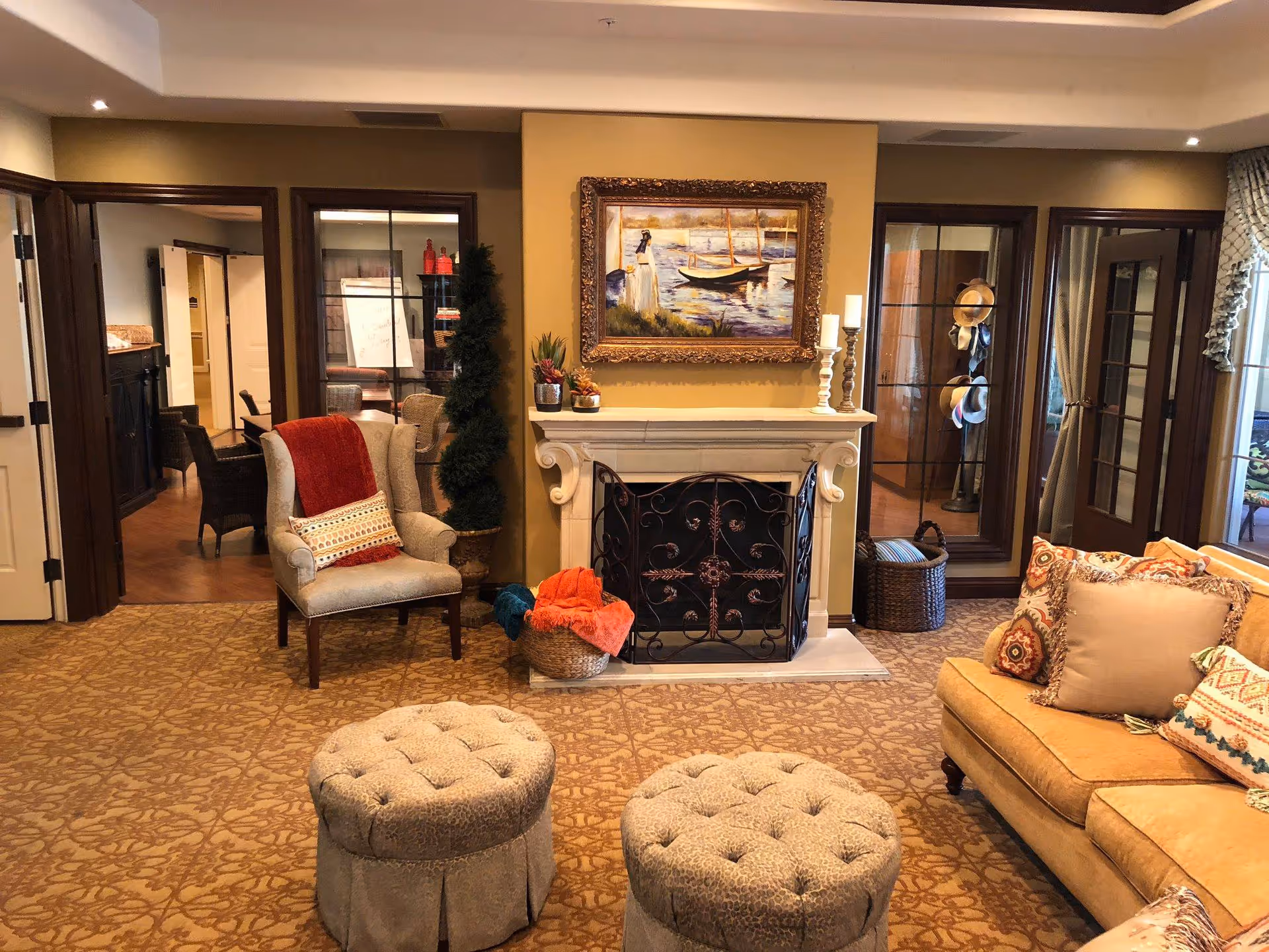 A cozy living room with a beige patterned carpet, a beige armchair with a red throw and decorative pillow, two round tufted ottomans, and a beige sofa with multiple patterned cushions. There is a white fireplace with a decorative black screen, topped with candles and small plants. Above the fireplace hangs a framed painting of boats on water. The room has warm yellow walls and wooden door frames leading to other rooms, with glass doors and windows showing additional seating areas and decor.