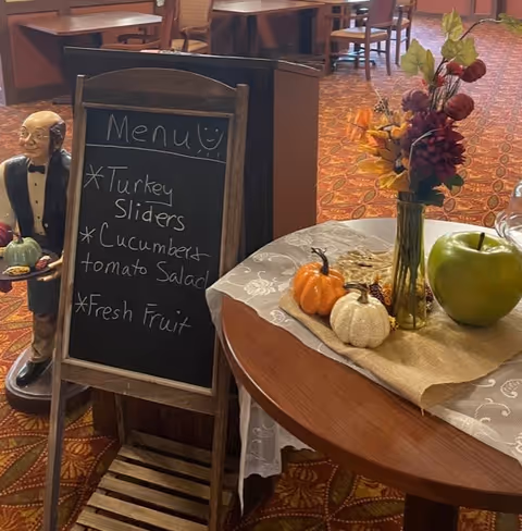 Chalkboard menu and a fall-themed table centerpiece with small pumpkins and flowers in a senior living dining room.