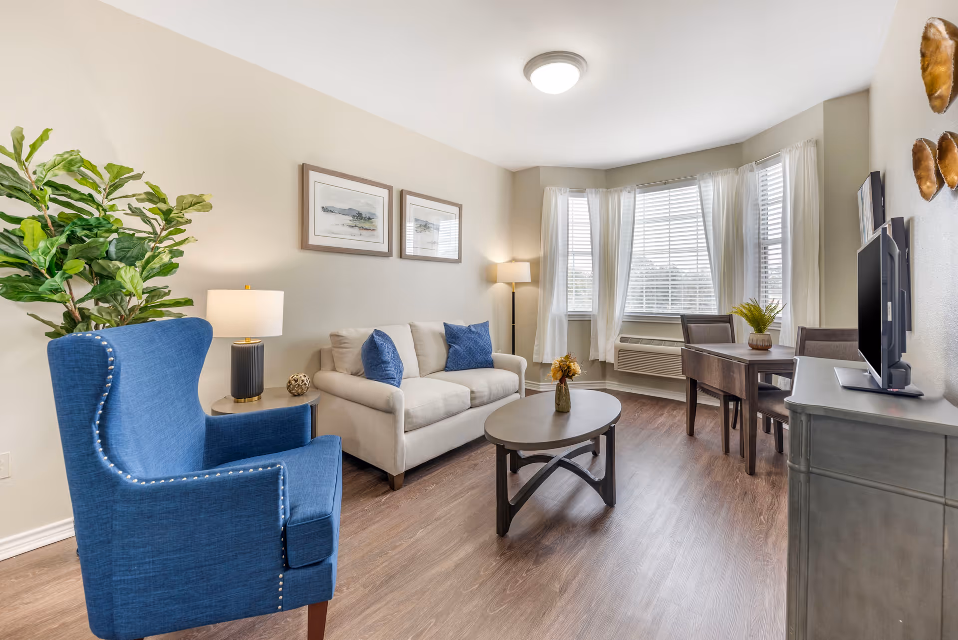 A bright living room with a beige sofa adorned with blue pillows, a blue armchair, a wooden coffee table with a small vase of flowers, a wooden dining table with two chairs near a bay window with white curtains, a TV on a gray cabinet, and decorative wall art.