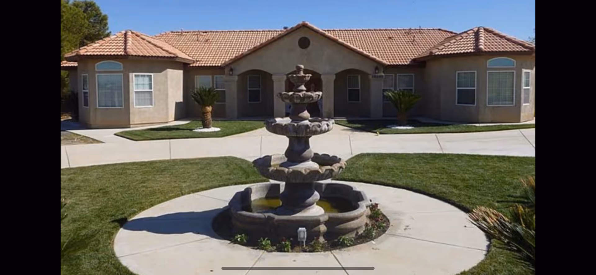 Exterior view of a single-story building with a tiled roof, featuring a central entrance with columns and two symmetrical wings. In front of the building, there is a circular concrete pathway surrounding a multi-tiered stone fountain, with green grass and small palm trees on either side.