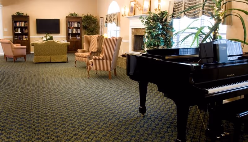 A spacious living room area with patterned carpet, a black grand piano on the right, two striped armchairs, a green sofa, a pink armchair, bookshelves, a flat-screen TV mounted on the wall, and a fireplace with plants and framed pictures above it.