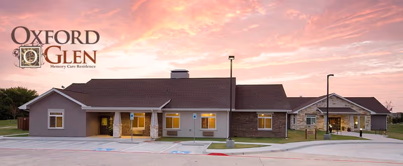 Exterior view of Oxford Glen Owasso, a memory care residence building, during sunset with a colorful sky. The building has a combination of brick and siding with multiple windows and a covered entrance. There are parking spaces and street lamps in front of the building.