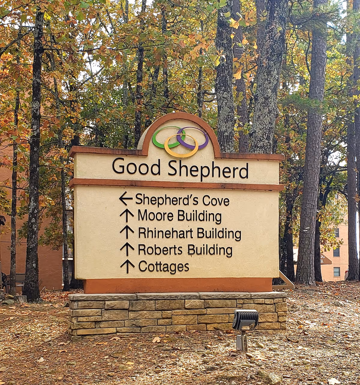 Outdoor sign for Good Shepherd Community surrounded by trees with autumn foliage. The sign directs to Shepherd's Cove, Moore Building, Rhinehart Building, Roberts Building, and Cottages. A spotlight is positioned in front of the sign.