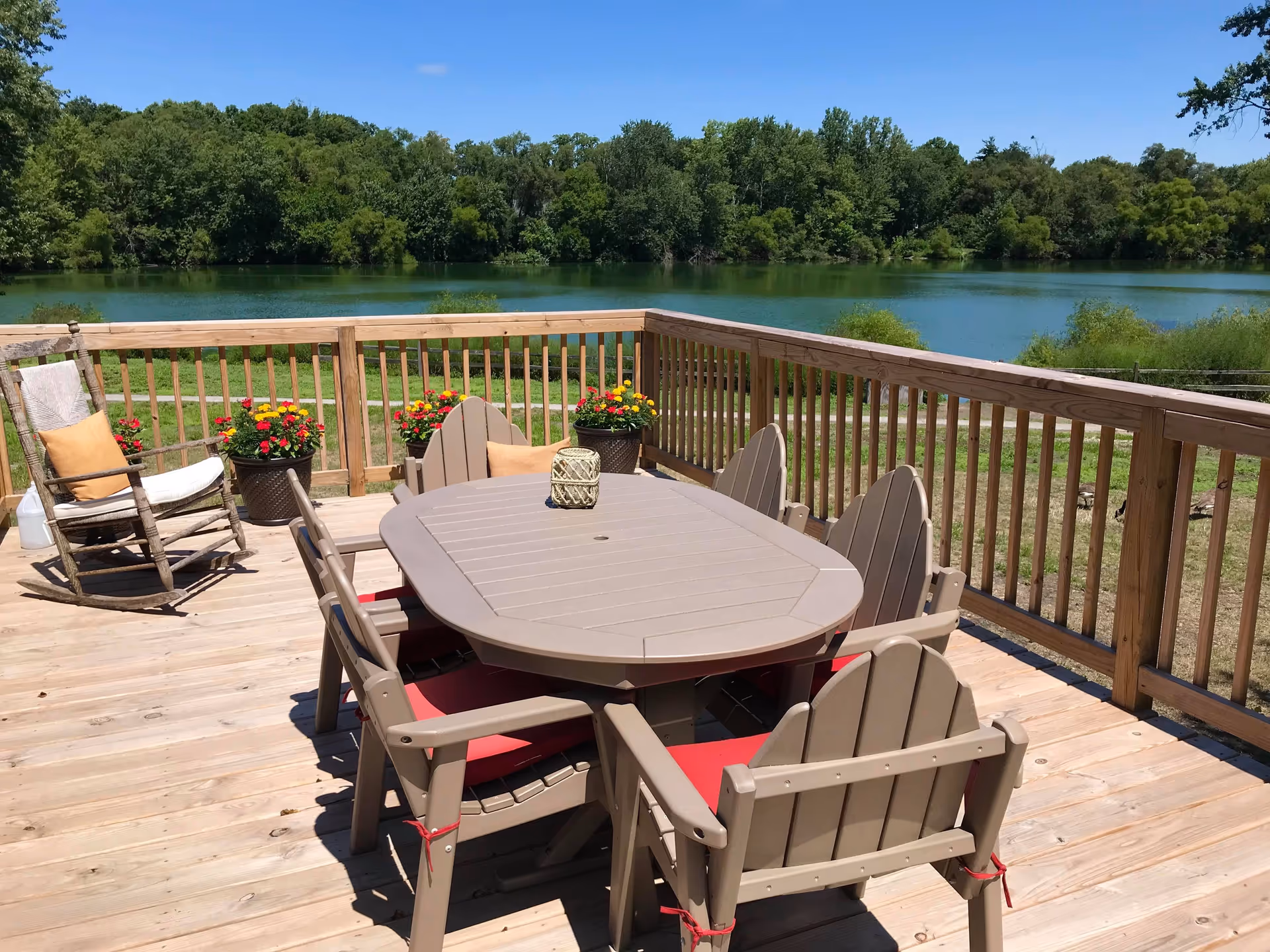 Outdoor wooden deck with a table and six chairs with red cushions, a rocking chair with a cushion and towel, potted flowers along the railing, overlooking a lake with trees in the background under a clear blue sky.