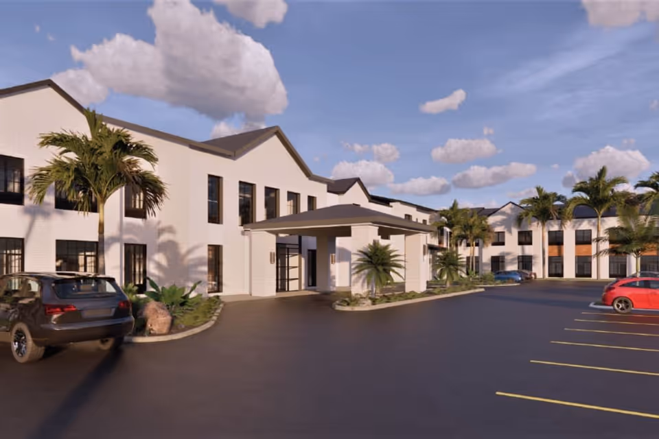 Exterior view of a two-story senior living facility building with white walls, large windows, and a covered entrance. Several palm trees and plants are around the building, and cars are parked in the parking lot under a partly cloudy sky.