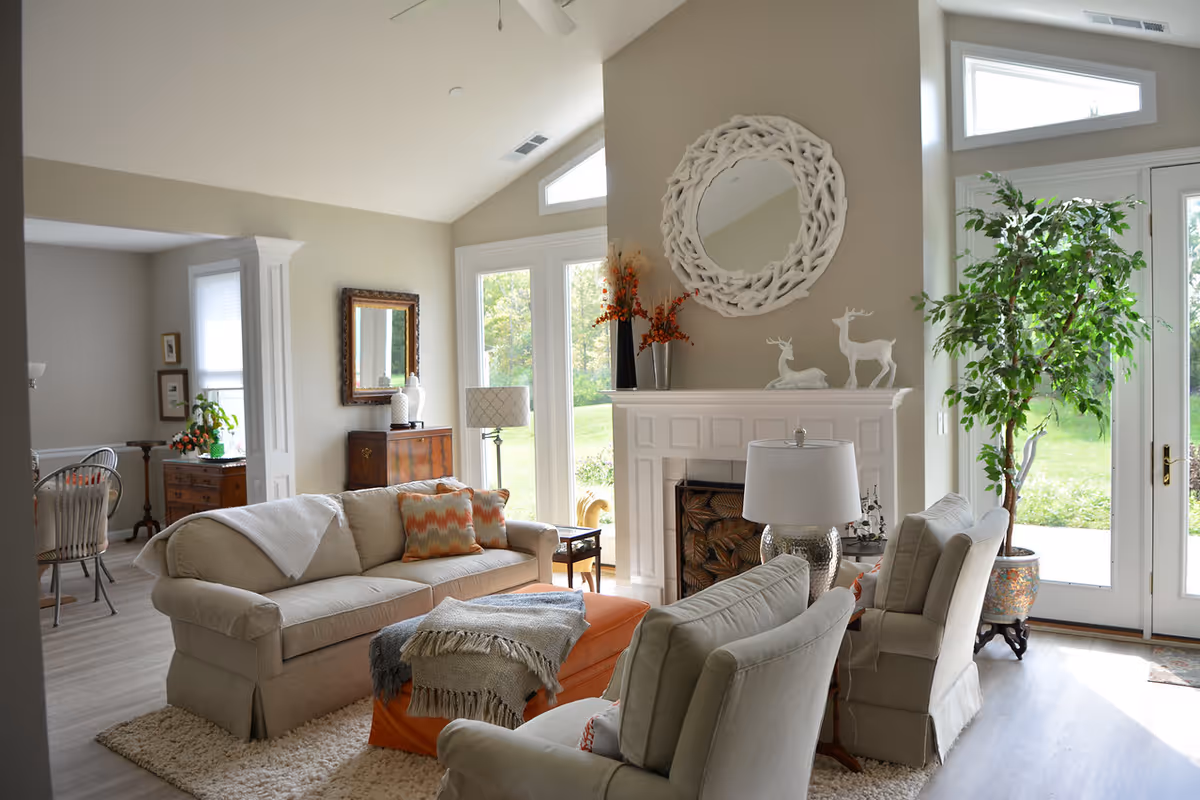 A bright and cozy living room with a beige sofa, two armchairs, and an orange ottoman. The room features a white fireplace with decorative deer figurines and a round white mirror above it. Large windows and glass doors let in natural light and offer a view of greenery outside. There is a tall potted plant near the door and a wooden cabinet with a lamp and mirror in the background.