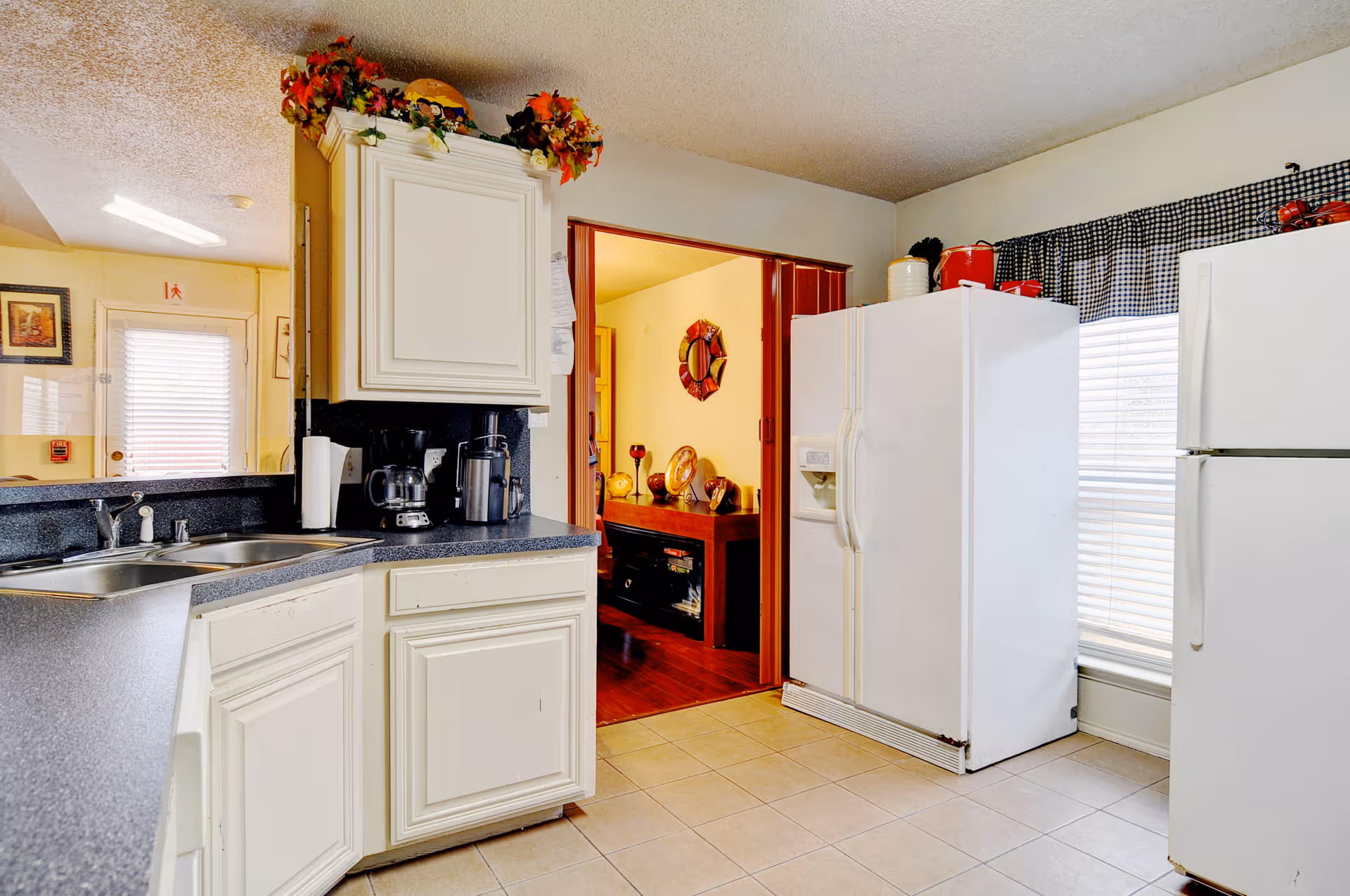 Bright kitchen with white cabinets, dual refrigerators, a sink and countertop appliances, and a view into an adjacent room.