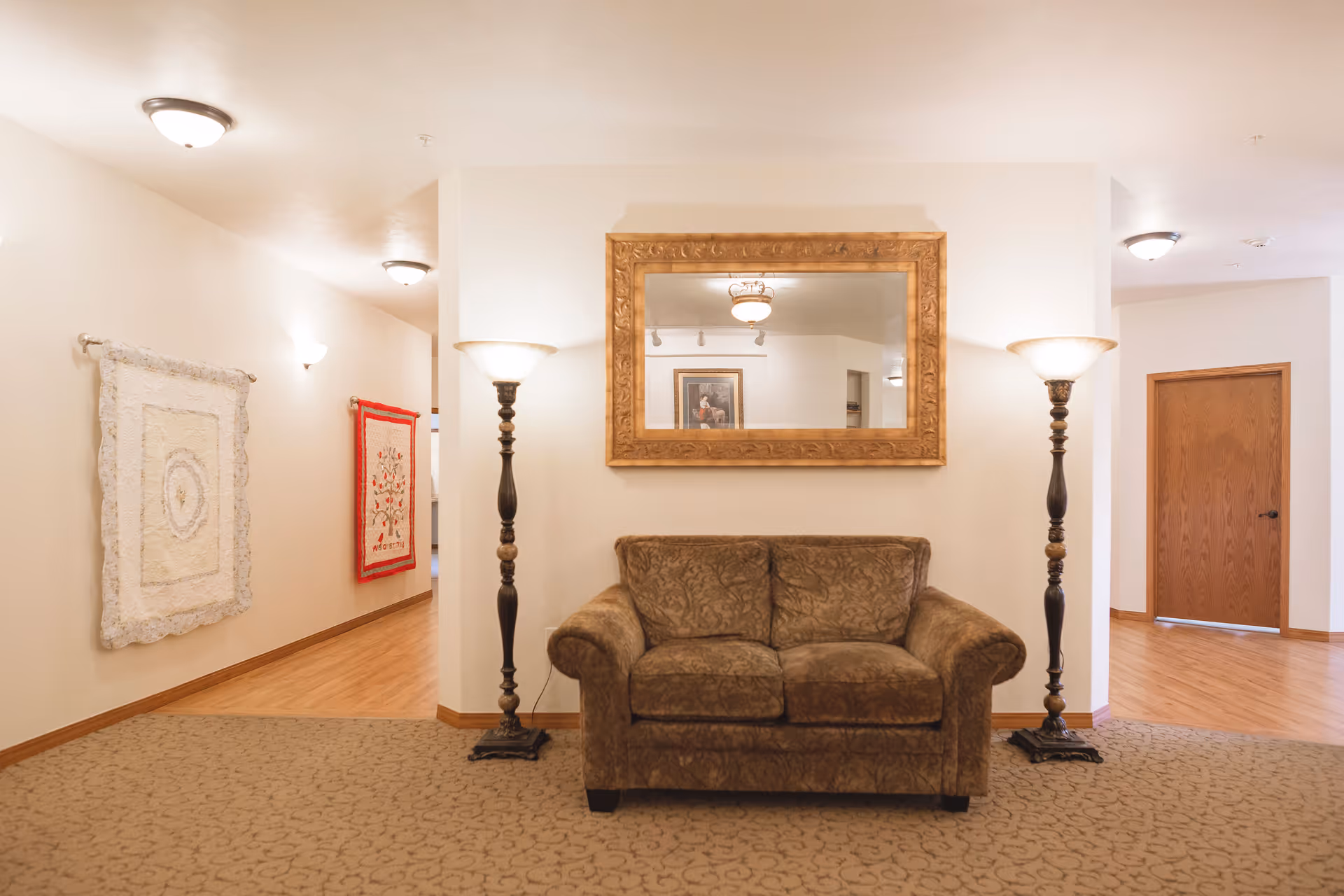 A cozy interior common area featuring a brown patterned loveseat centered against a white wall. Above the loveseat is a large ornate gold-framed mirror. On either side of the loveseat are tall floor lamps with frosted glass shades. The floor is carpeted in front of the loveseat and transitions to wood flooring in the adjacent hallway. The hallway walls display two decorative quilts. A wooden door is visible in the background.