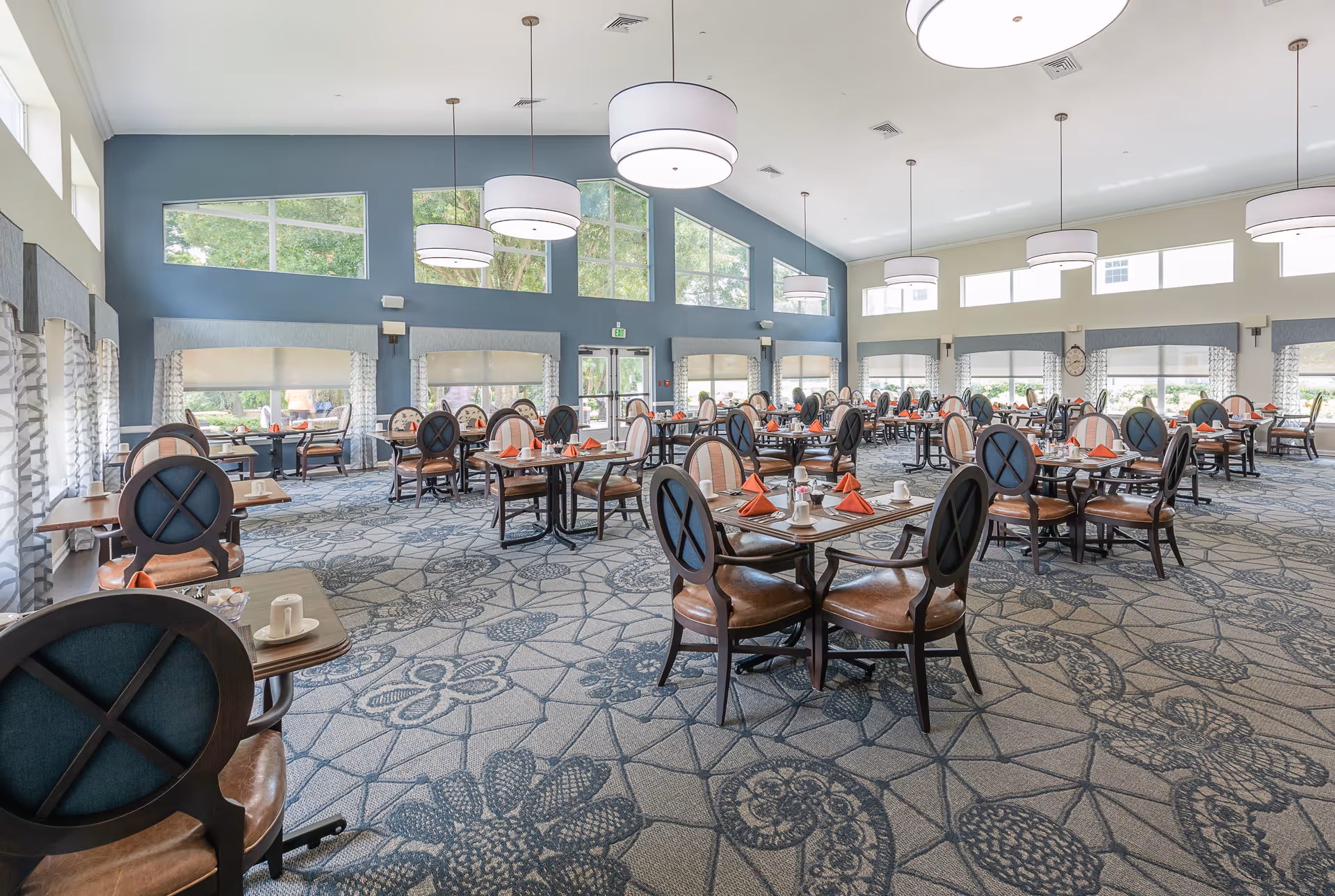 Spacious dining room with multiple tables and chairs arranged neatly. Each table is set with white cups, plates, and orange folded napkins. Large windows and high ceilings with modern pendant lights provide ample natural light. The carpet has a floral pattern, and the walls are painted blue and white.
