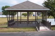 A wooden gazebo with a shingled roof situated on a grassy area near a small body of water. The gazebo has railings and a ramp leading up to it, with trees and a parking lot visible in the background.