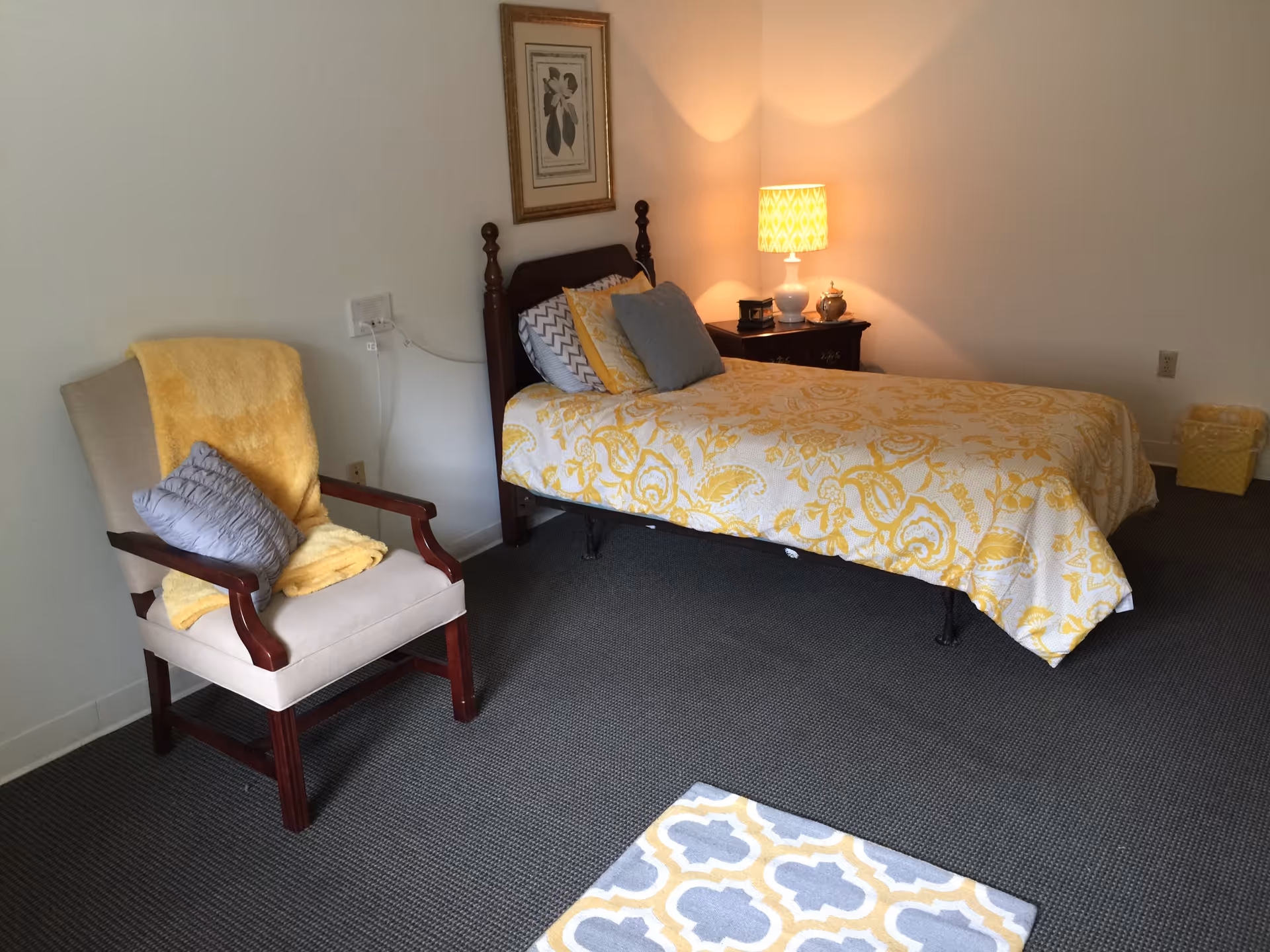 A bedroom with a single bed covered in a yellow and white patterned bedspread, several pillows, a wooden nightstand with a lit lamp, and a framed picture on the wall. There is also an upholstered chair with a yellow blanket and a gray pillow, a small patterned rug on the floor, and a yellow wastebasket in the corner.