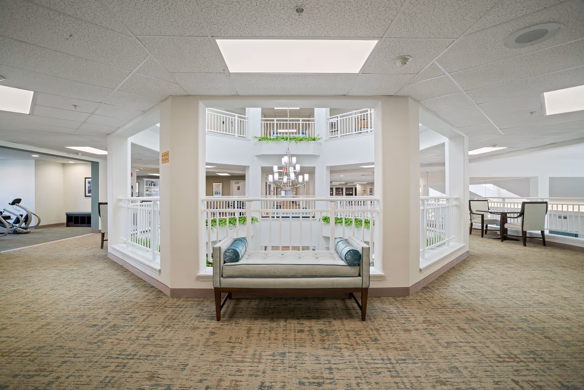 Interior view of a senior living facility with a cushioned bench in the foreground, overlooking a central atrium with white railings and green plants. The area is well-lit with ceiling lights and has carpeted floors. There are chairs and tables visible on the right side and exercise equipment on the left side.
