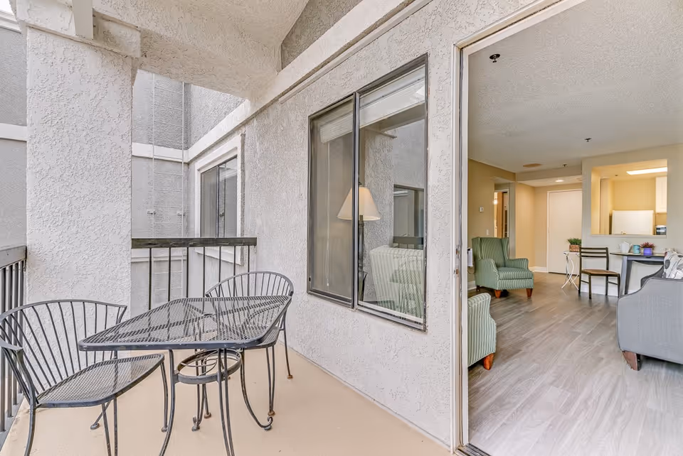 Outdoor balcony area with a black metal table and two matching chairs. The balcony has beige textured walls and a railing. Through a sliding glass door, an interior living space is visible with green armchairs, a gray sofa, a small dining table with chairs, and a kitchen area in the background.
