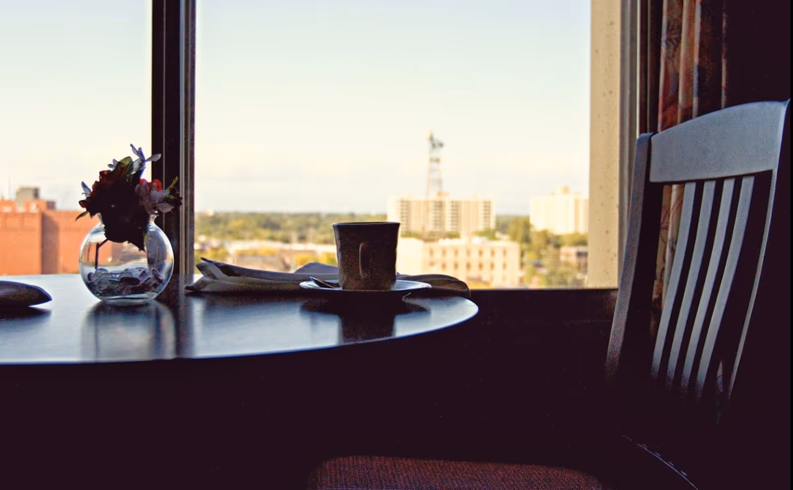 A small round table by a window holding a glass vase with flowers and a cup on a saucer with a city view outside.