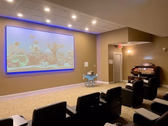 A cozy senior living facility room with several black cushioned chairs arranged in rows facing a large screen displaying an underwater scene with fish and coral. The room has beige walls and carpet, recessed ceiling lights, a small cart with items on it, and a vintage-style organ in the corner.