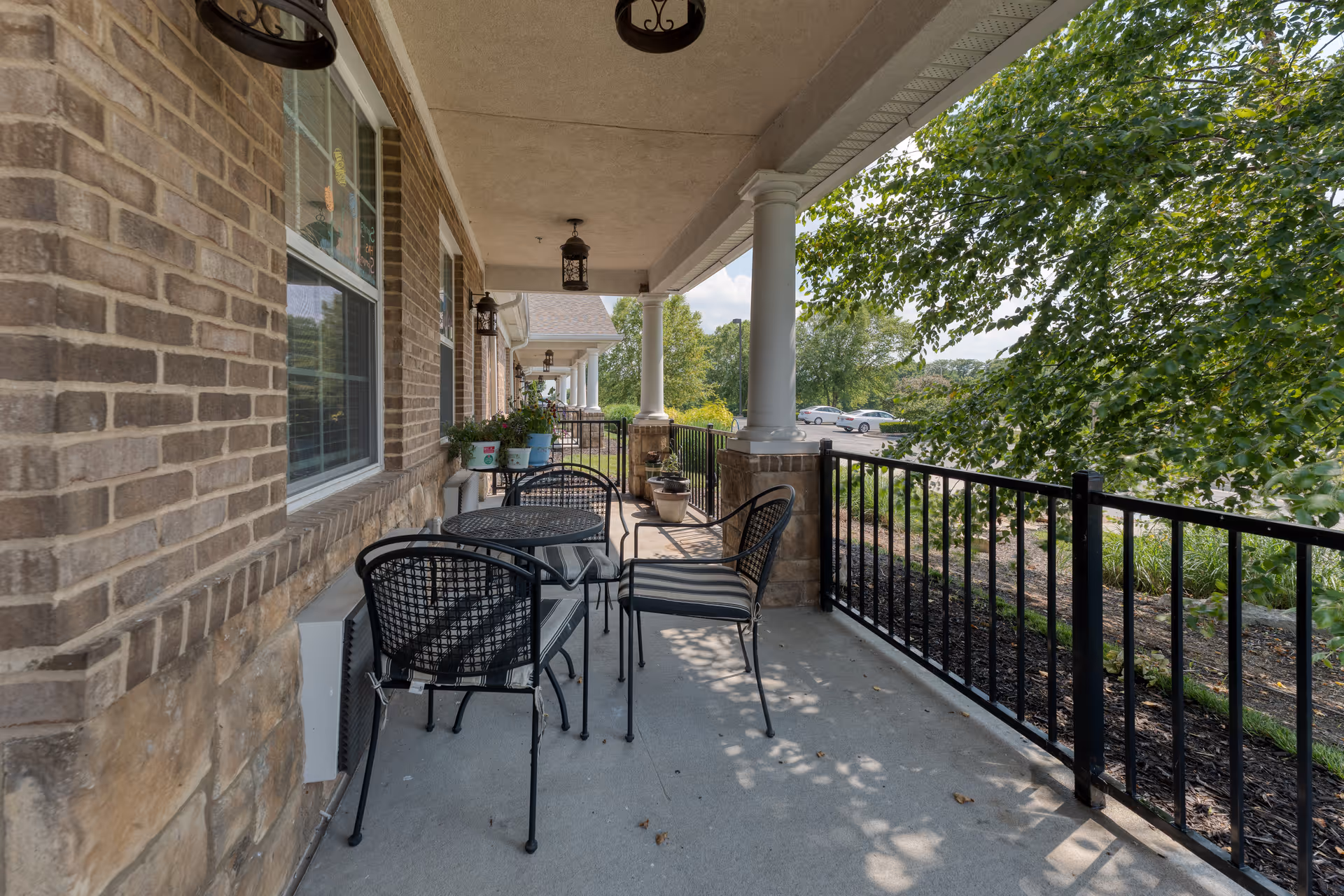 Covered outdoor patio area with black metal tables and chairs, brick walls, white columns, and a black metal railing. There are potted plants on the tables and ledge, with trees and a parking lot visible in the background.