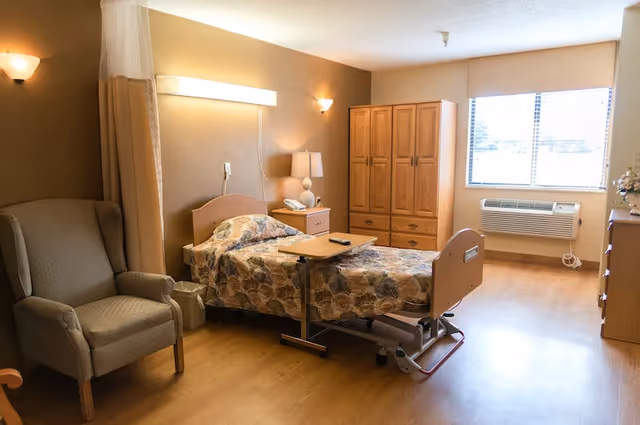 A well-lit senior living bedroom featuring a single hospital-style bed with floral bedding, an adjustable overbed table, a comfortable armchair, a wooden wardrobe, a nightstand with a lamp and telephone, a window with blinds, and a wall-mounted air conditioning unit.
