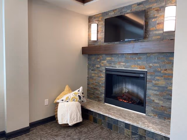 Cozy corner of a living room featuring a modern electric fireplace set in a stone wall with a mounted flat-screen TV above it. A basket with decorative pillows and a white throw blanket is placed on the floor next to the fireplace. The room has neutral-colored walls and carpeted flooring.