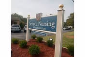 A freestanding blue sign that reads 'Seneca Nursing & Rehabilitation' set in a landscaped bed with a building and parked cars in the background.