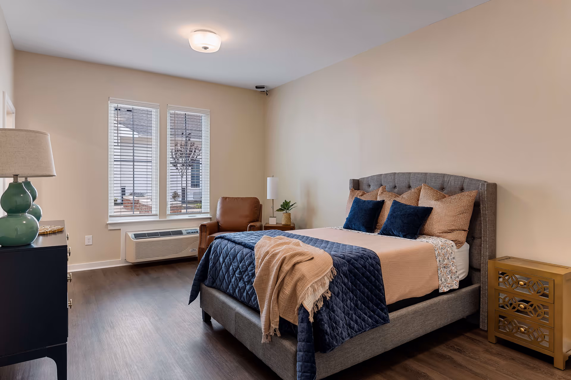 A neatly arranged bedroom with a gray upholstered bed featuring multiple pillows and a blue quilted blanket. There is a beige throw draped over the bed. Next to the bed is a gold-colored nightstand with decorative drawer fronts. A brown leather armchair and a small side table with a lamp and plant are positioned near two windows with white blinds. A black dresser with a green lamp is visible on the left side of the room. The floor is wooden, and the walls are painted beige.
