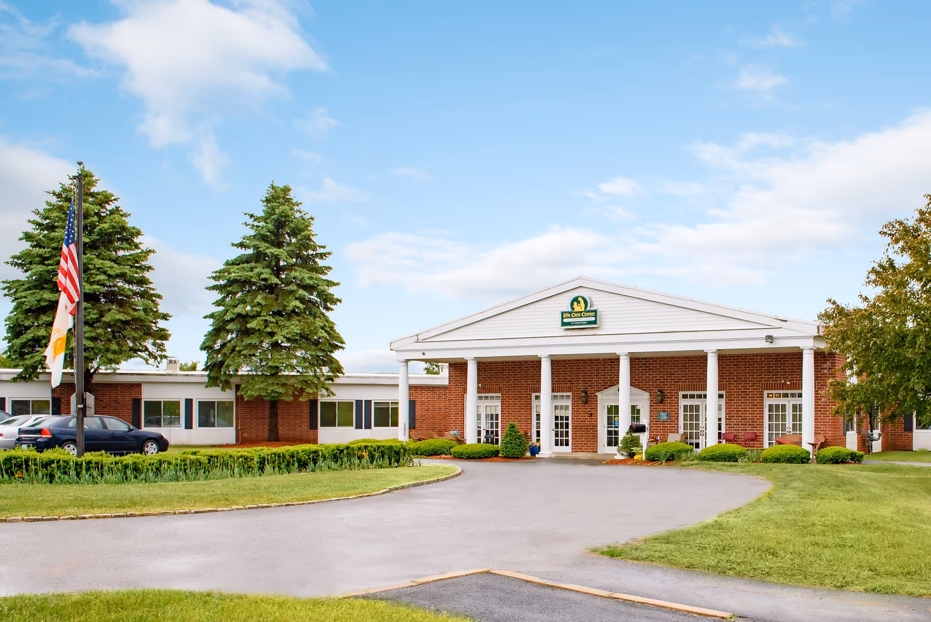 Front exterior view of Life Care Center of Nashoba Valley, a single-story brick building with white columns at the entrance, surrounded by green grass, trees, and a driveway. Two flags are flying on flagpoles to the left of the building under a partly cloudy sky.