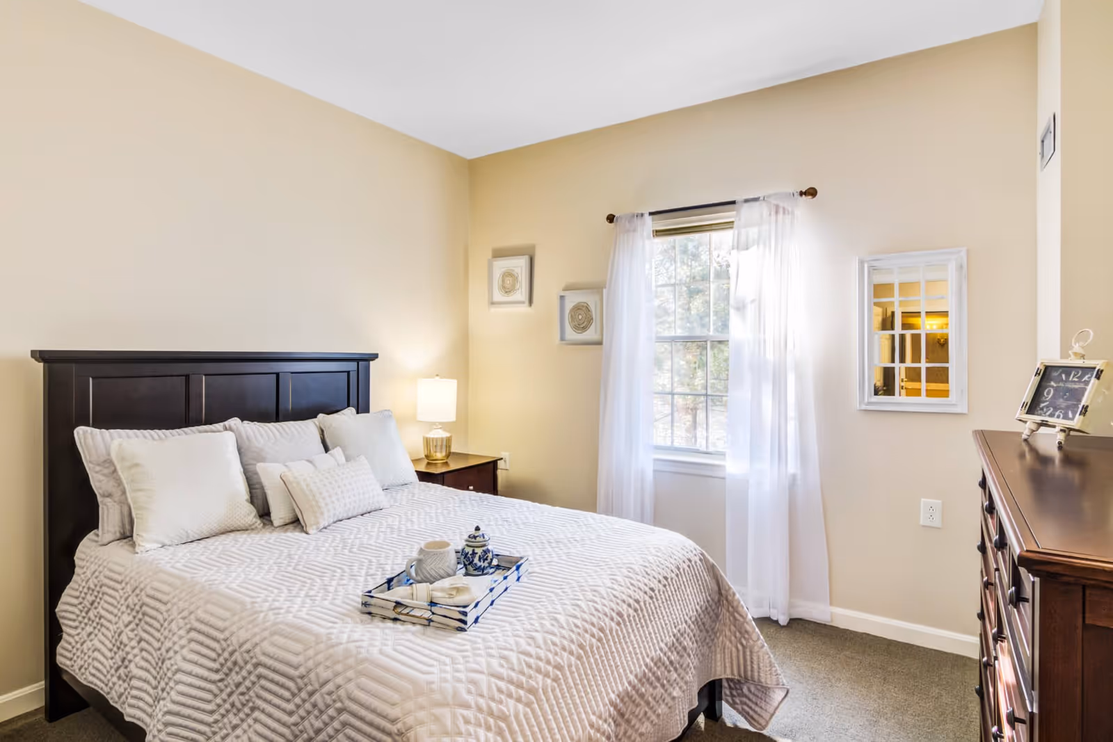 A bright bedroom with a large bed featuring a dark wooden headboard and white patterned bedding. There are several pillows on the bed and a tray with a teapot and cup. A nightstand with a lamp is beside the bed. A window with sheer white curtains lets in natural light. A dresser with a small clock and a wall mirror are also visible.