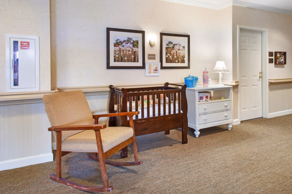 Hallway interior with a wooden rocking chair, a small wooden crib and a white dresser with a lamp against a wallpapered wall.