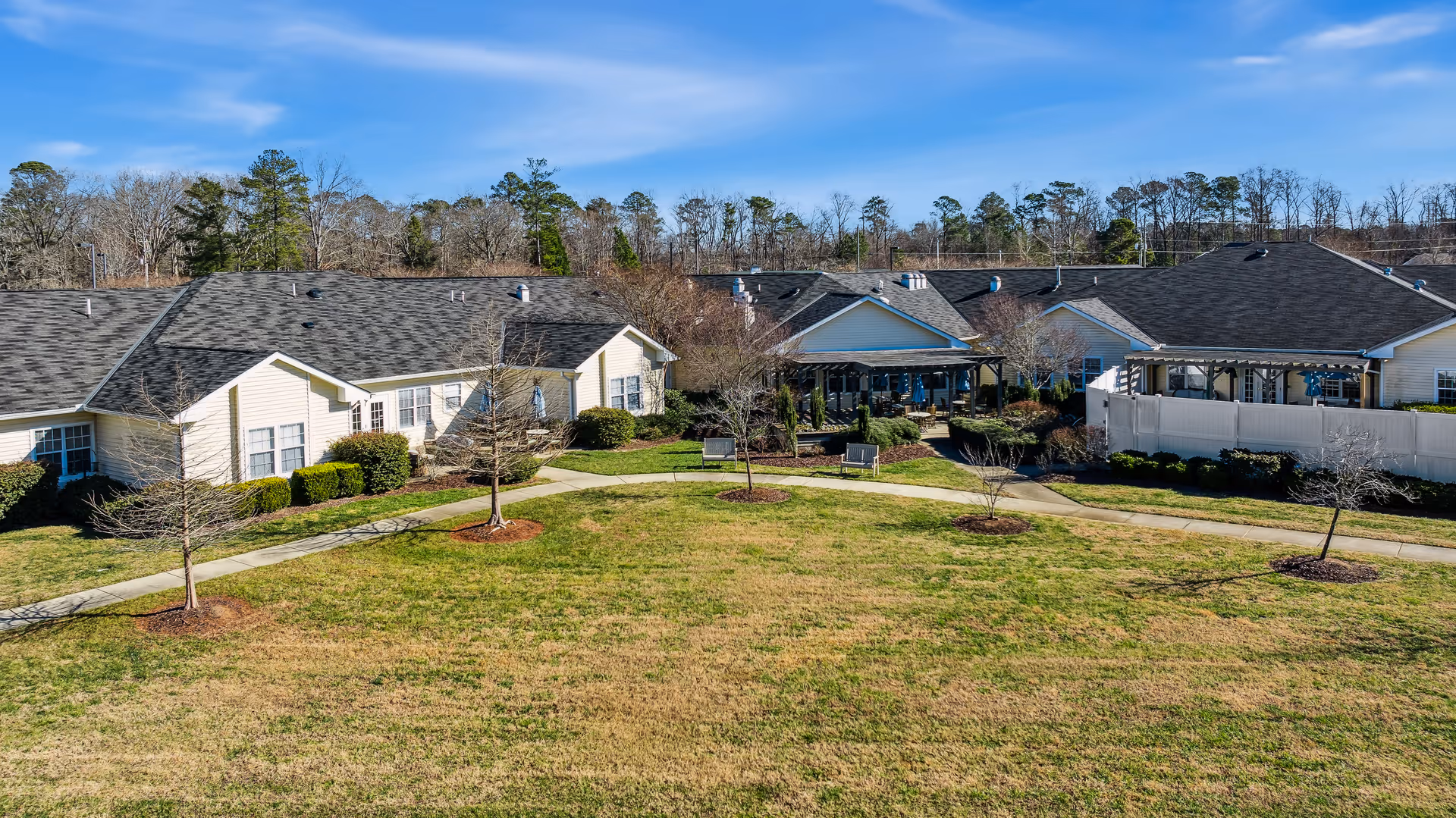 Low-rise beige residential buildings surround a grassy courtyard with walkways and small trees under a blue sky.