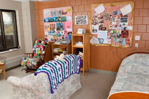 A resident room with a bed, patchwork chair, and collage boards on an orange brick wall.