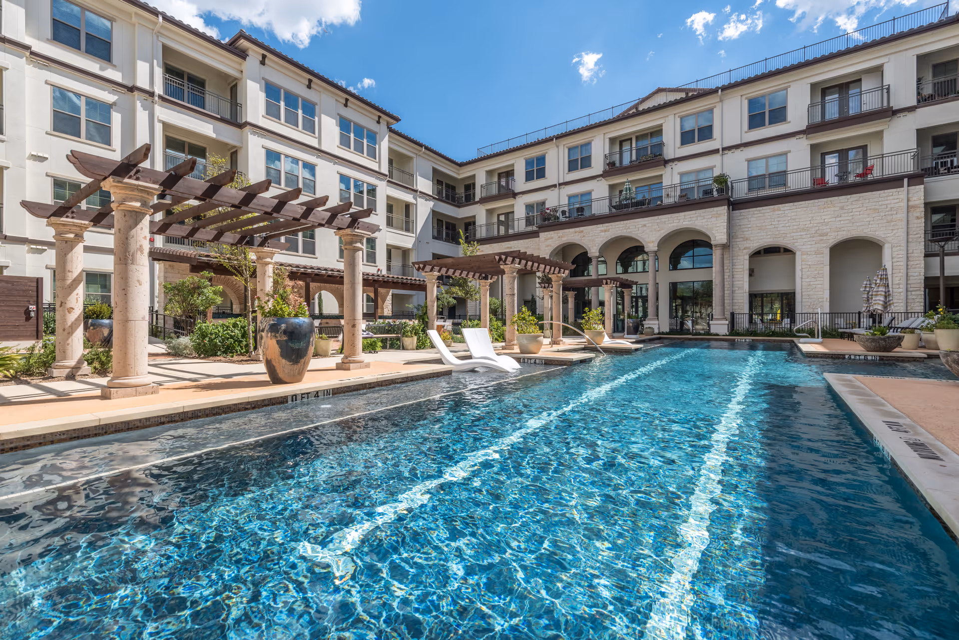 Outdoor swimming pool with clear blue water in front of a multi-story senior living facility building. The pool area features lounge chairs partially submerged in the water, large decorative planters, and pergolas with stone columns. The building has balconies and large arched windows under a bright blue sky with some clouds.
