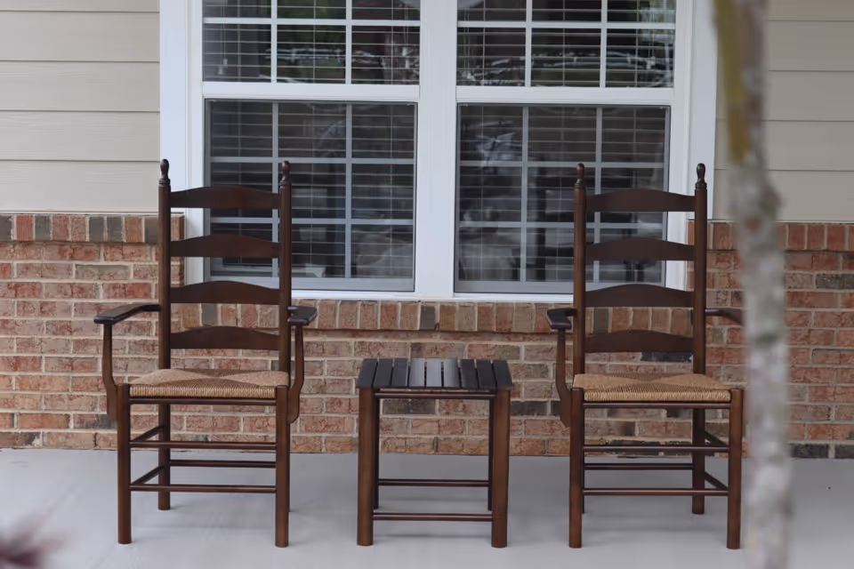 Two wooden chairs with woven seats and a small wooden table between them are placed on a concrete porch in front of a window with white blinds. The exterior wall is made of beige siding and red brick.