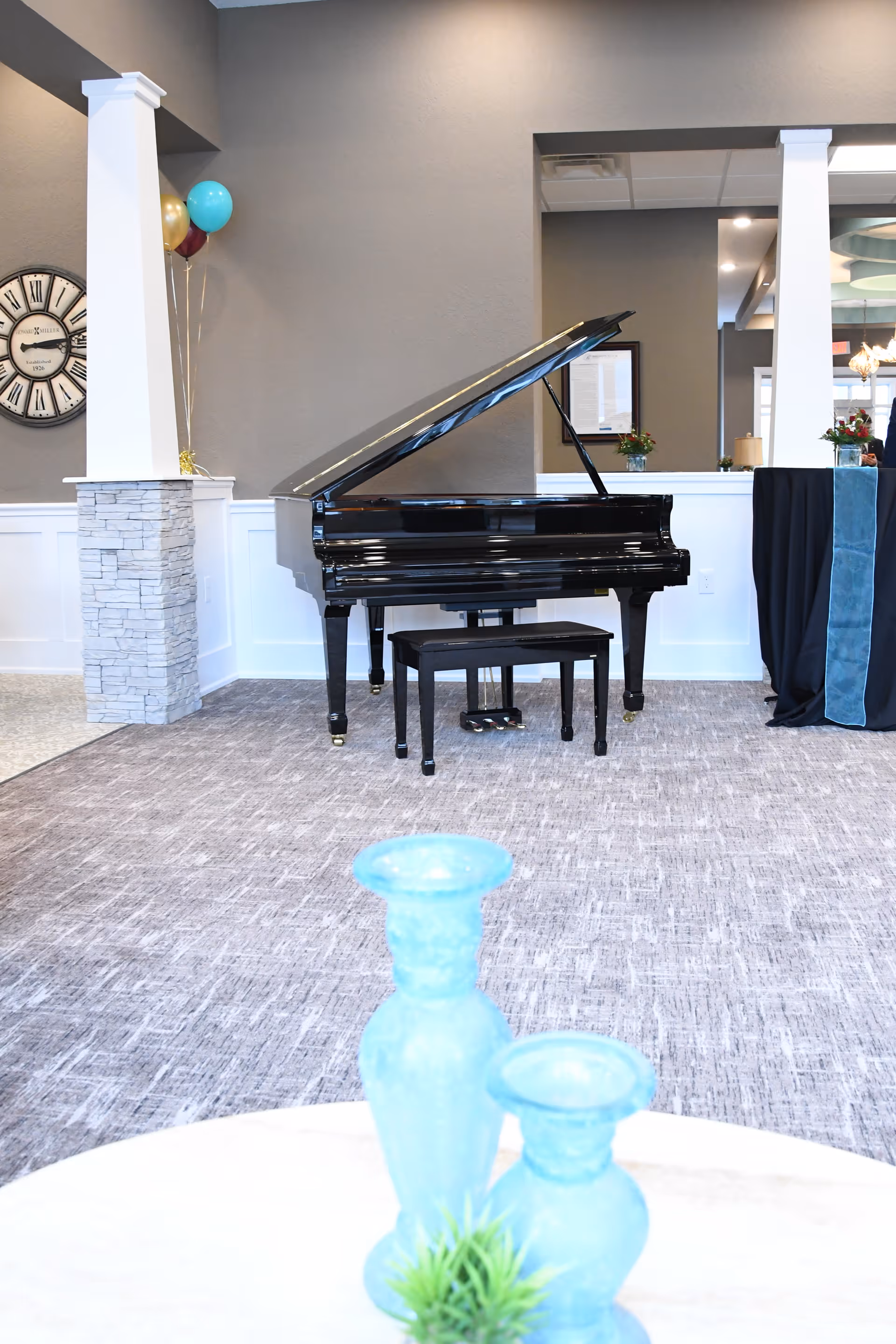 Interior view of a senior living facility featuring a black grand piano with a matching bench on a carpeted floor. The background includes a gray wall with white wainscoting, a large decorative clock, and some balloons. In the foreground, there are two blue glass vases and a small green plant on a white table.