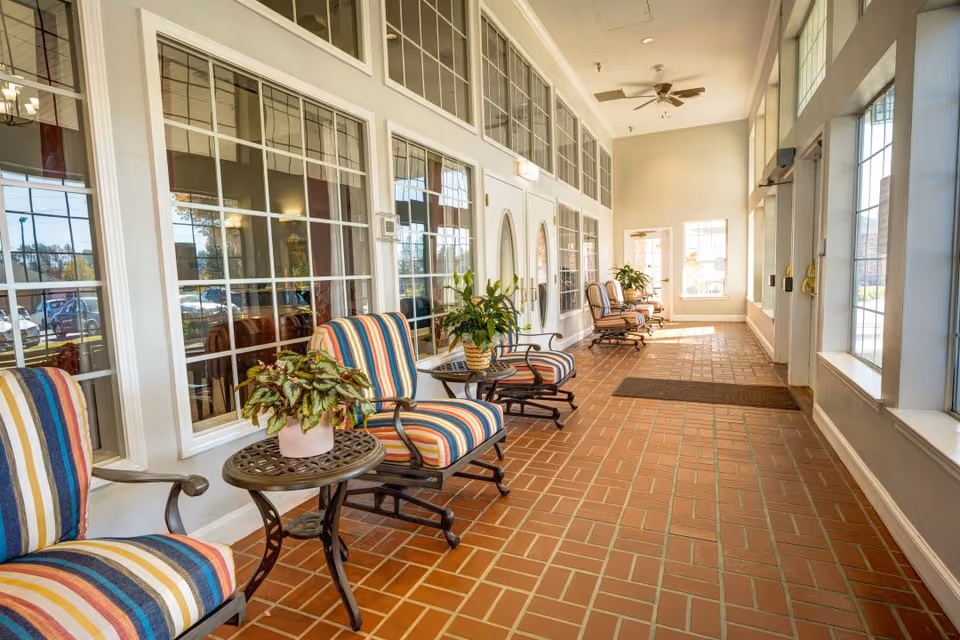 A bright indoor hallway with large windows on both sides, featuring several striped cushioned chairs and small round tables with potted plants. The floor is covered with reddish-brown tiles, and ceiling fans are visible above.