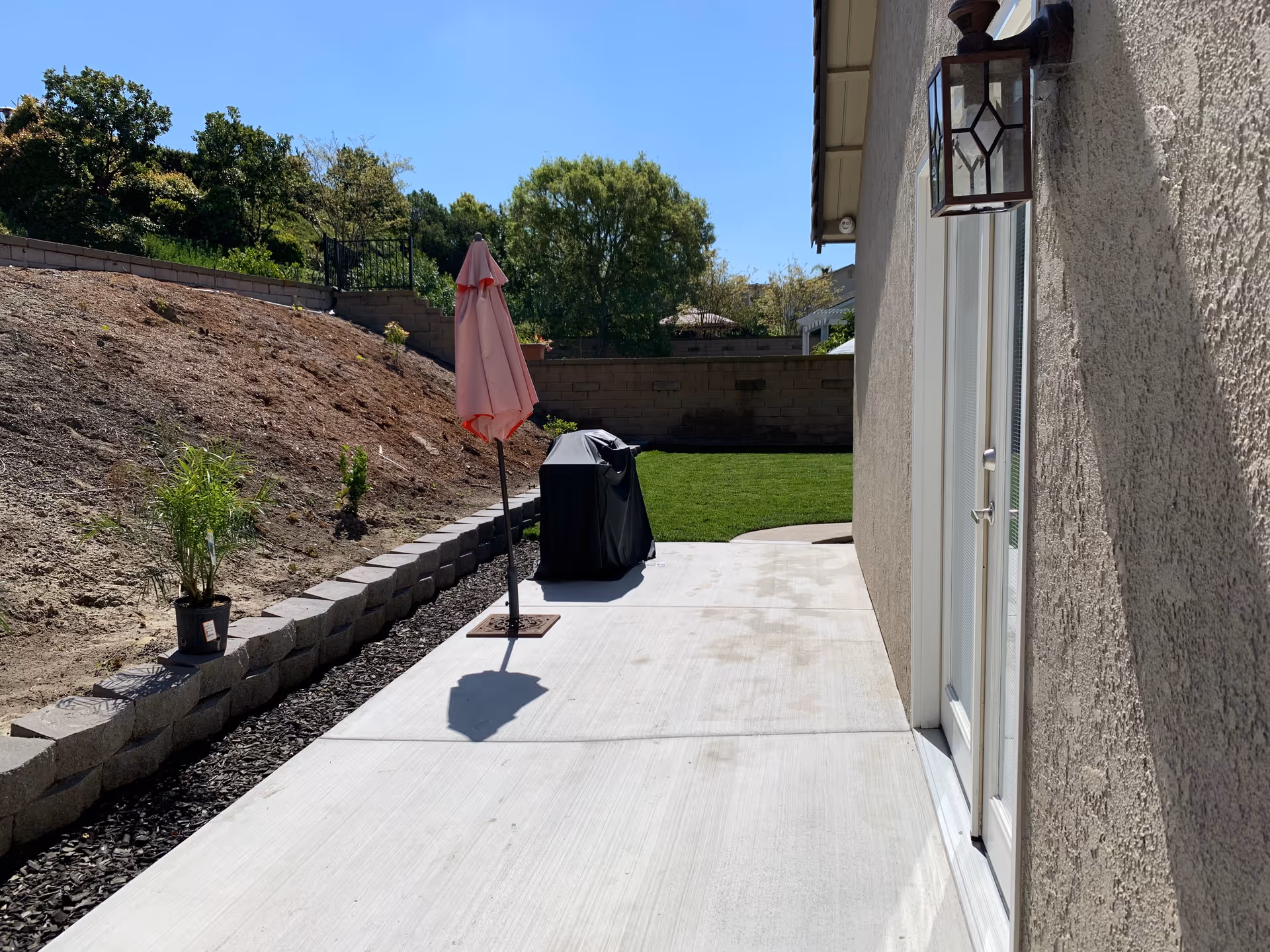 Outdoor patio area with a concrete walkway, a closed pink umbrella, a covered barbecue grill, a small potted plant on a retaining wall, and a grassy lawn bordered by a brick wall under a clear blue sky.