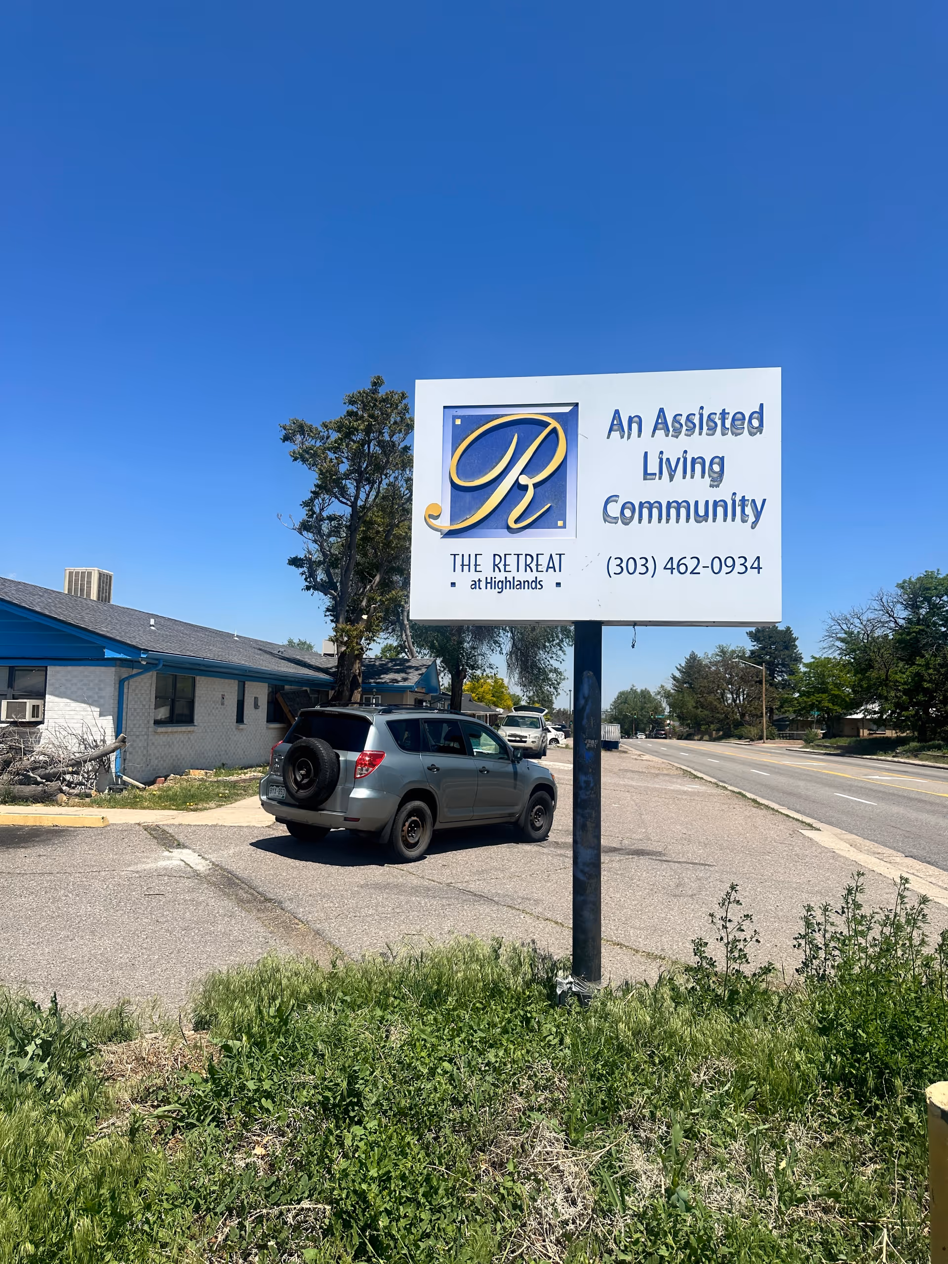 Outdoor view of a sign for The Retreat at Highlands, an assisted living community, with a parked silver SUV and a building with a blue roof in the background under a clear blue sky.