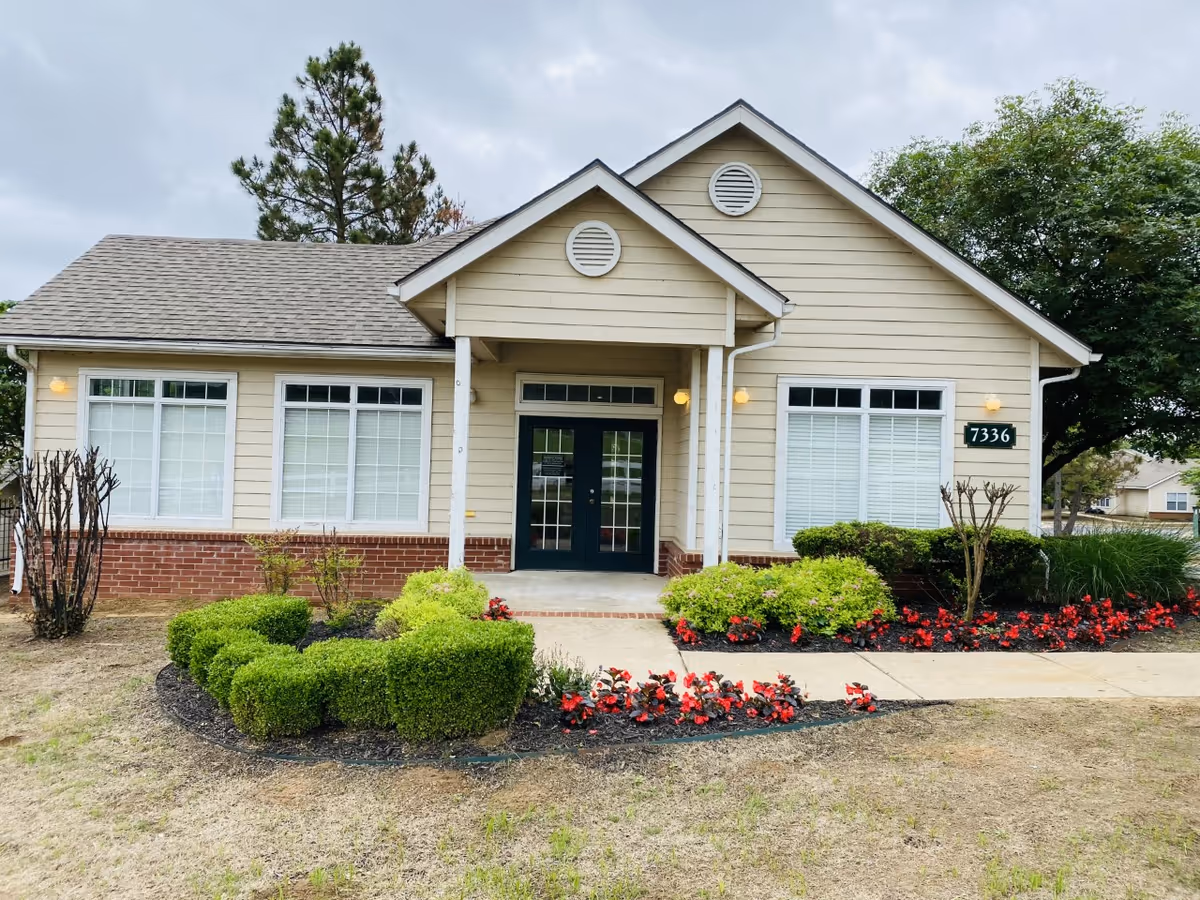 Exterior view of a single-story beige building with a gabled roof and white trim. The building has three large windows with white blinds and a double glass door entrance. There are neatly trimmed bushes and red flowers planted in front, along with a concrete walkway leading to the door. The building number 7336 is displayed on the right side. Trees and a cloudy sky are visible in the background.