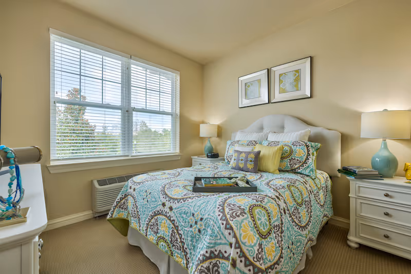 A bright bedroom with a large window covered by white blinds. The bed has a patterned bedspread in shades of teal, yellow, and brown, with multiple decorative pillows. There are two white nightstands on either side of the bed, each with a blue lamp and some books. Two framed artworks hang above the bed. The room has beige walls and carpeted flooring.
