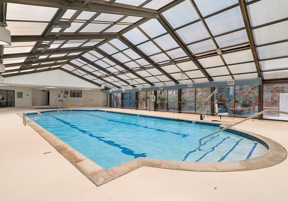 Indoor swimming pool with clear blue water under a large glass roof. The pool area has handrails, steps leading into the water, and a pool lift for accessibility. Large windows line one side of the pool area, showing some outdoor greenery and murals.