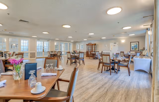 Interior view of the dining room at American House Hendersonville, featuring wooden tables set with dishes and flowers, large windows allowing natural light, and a cozy atmosphere.