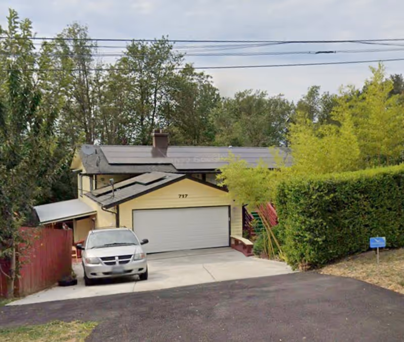 A two-story house with a light yellow exterior and a white garage door numbered 717. A silver car is parked in the driveway. The house is surrounded by trees and bushes, with a red fence on the left side and a green hedge on the right. The sky is partly cloudy.