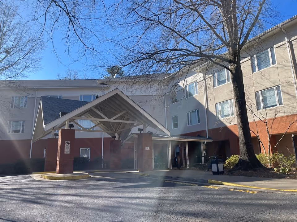 Covered entrance and canopy of a multi-story senior living building with a brick lower facade and leafless trees in front.