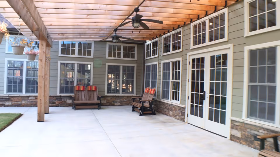 Covered outdoor patio area with wooden pergola ceiling, ceiling fans, two wooden benches with red and orange cushions, large windows, and glass doors leading inside a building.